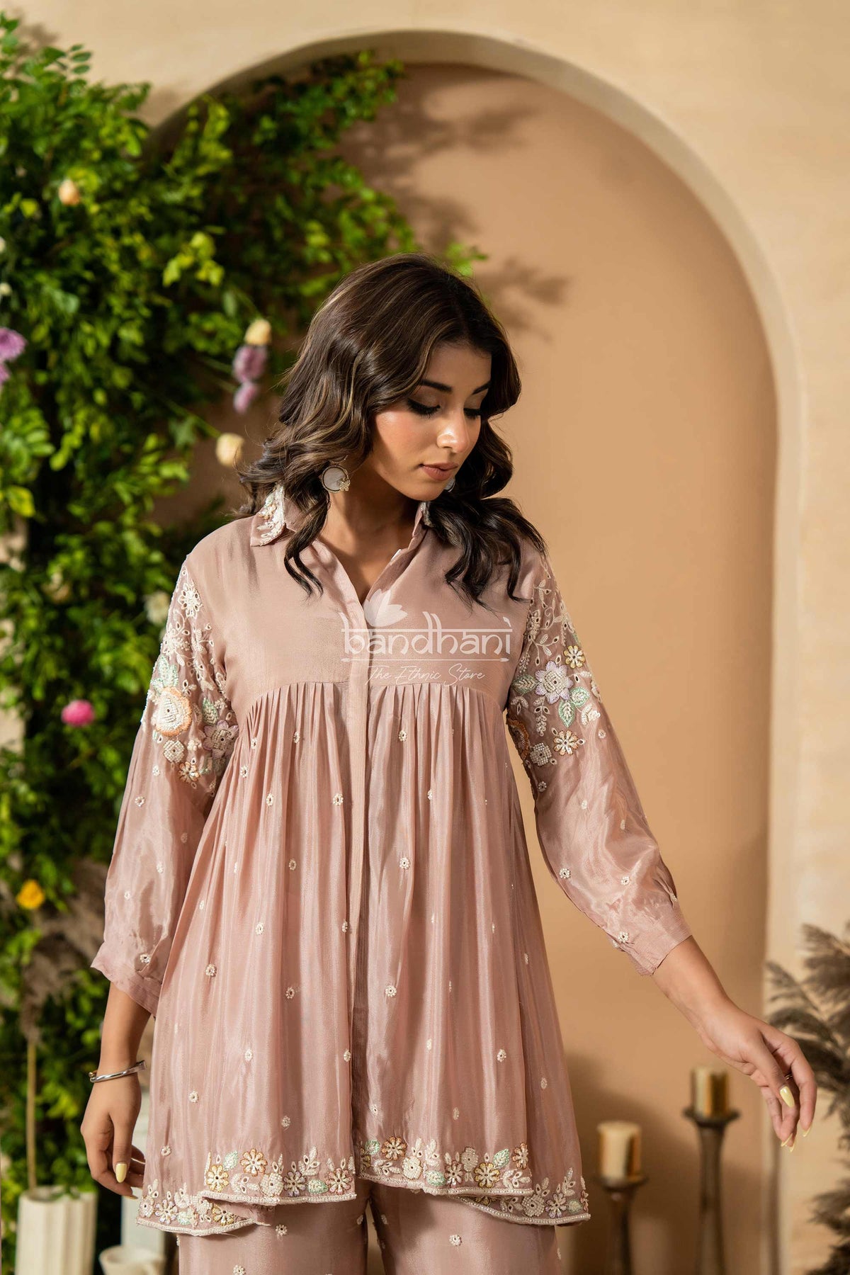 Woman wearing a light pink traditional outfit with intricate designs, standing against a decorative archway.