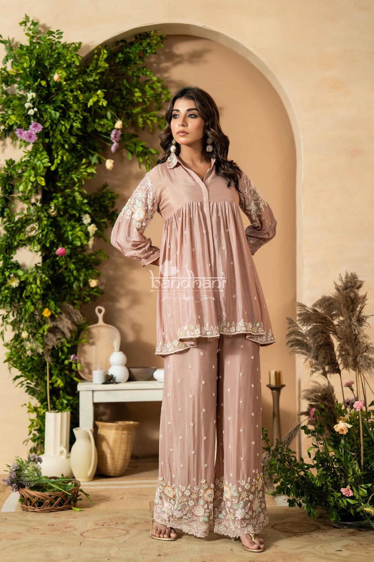 Woman in a pink traditional outfit standing in a decorated indoor setting with plants and decor.