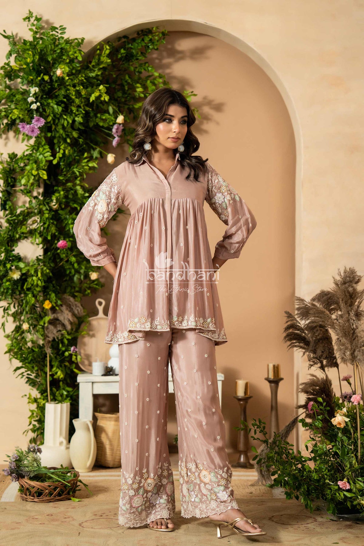 Woman in a pink embroidered outfit standing in a decorated room with plants and candles.
