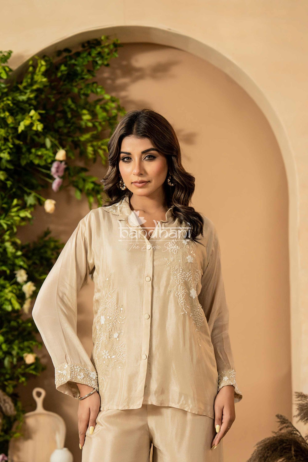 Woman wearing a beige embroidered outfit standing in front of an archway with greenery.