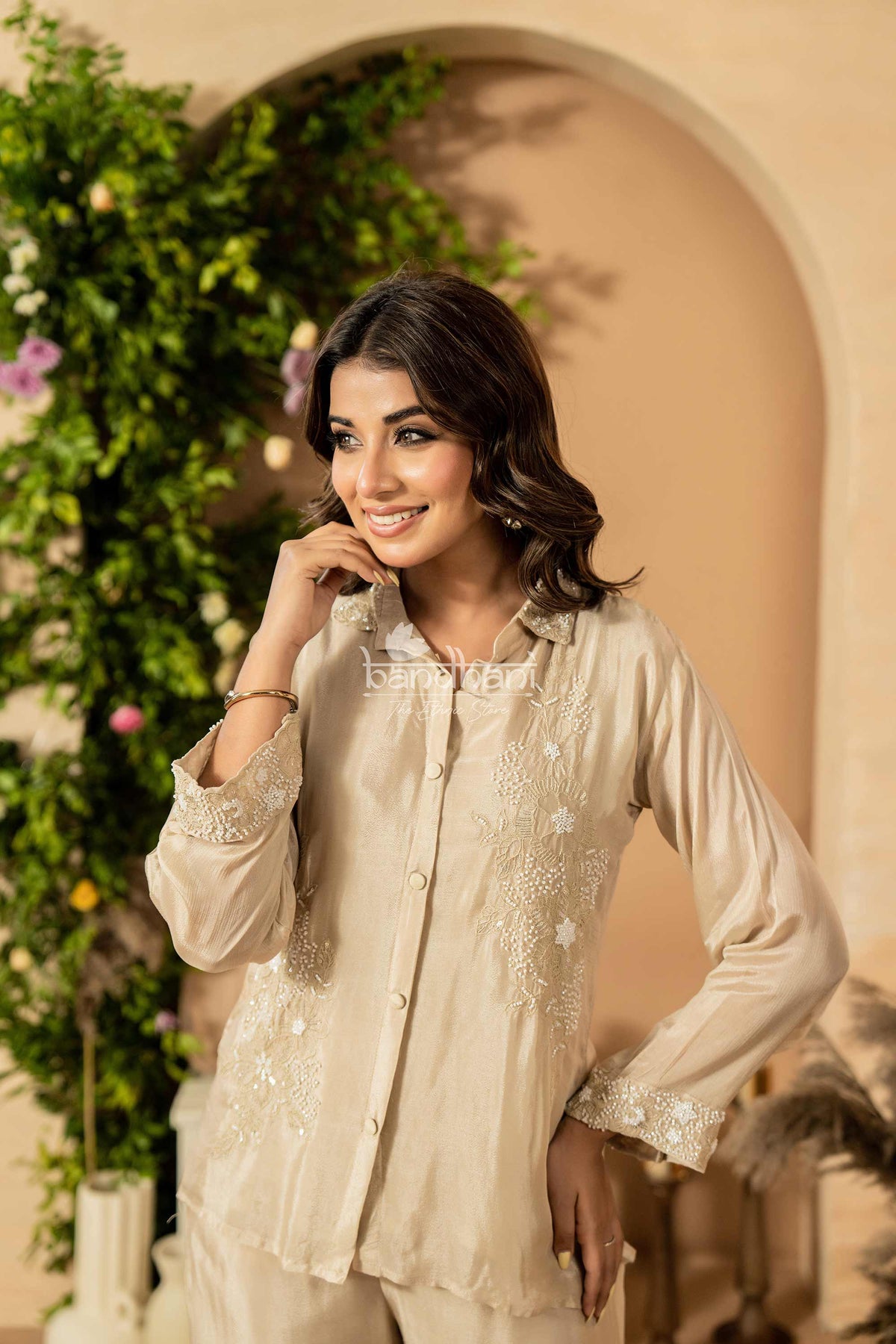 Woman in a beige embroidered outfit standing in front of a decorative archway with greenery.