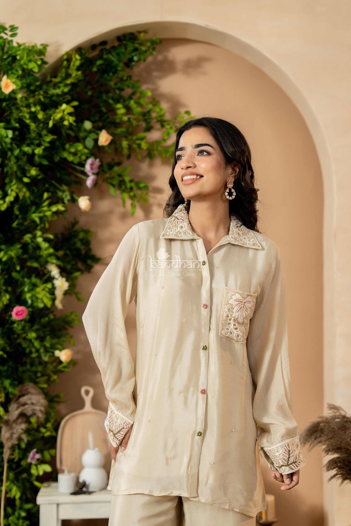 Woman wearing a beige embroidered shirt in an outdoor setting with greenery and flowers.