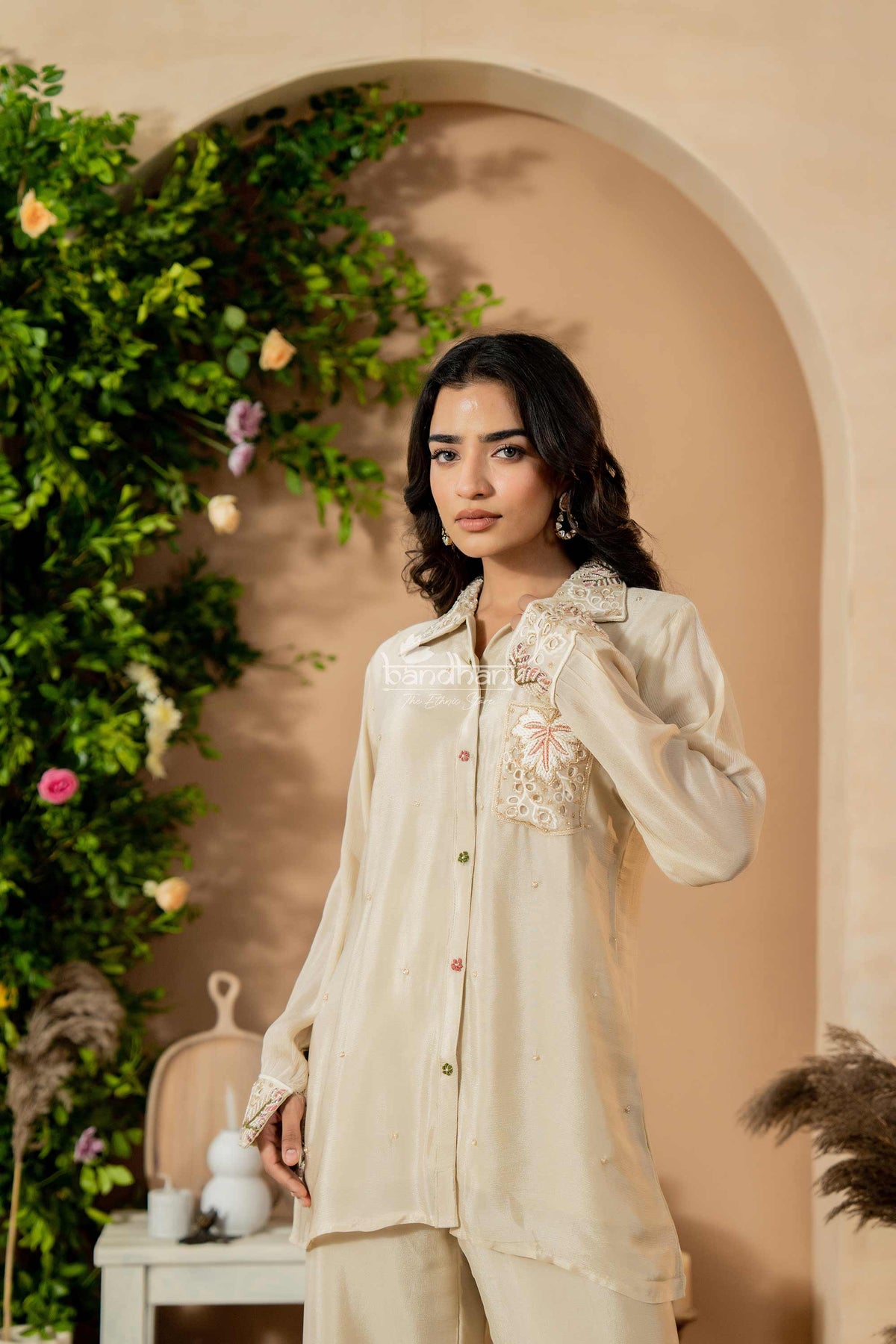 Woman wearing a beige embroidered shirt and pants in a decorated indoor setting with greenery.