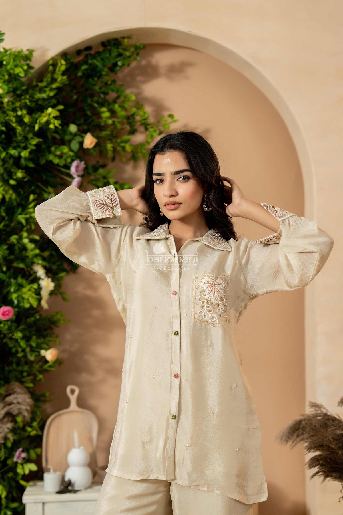Woman wearing a beige embroidered blouse in a decorative setting with plants and a beige wall.