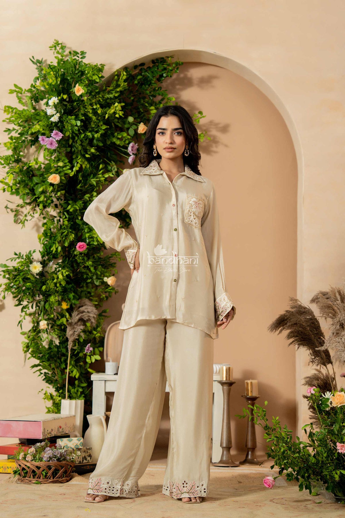 Woman in a beige outfit standing in a decorated indoor setting with plants and flowers.