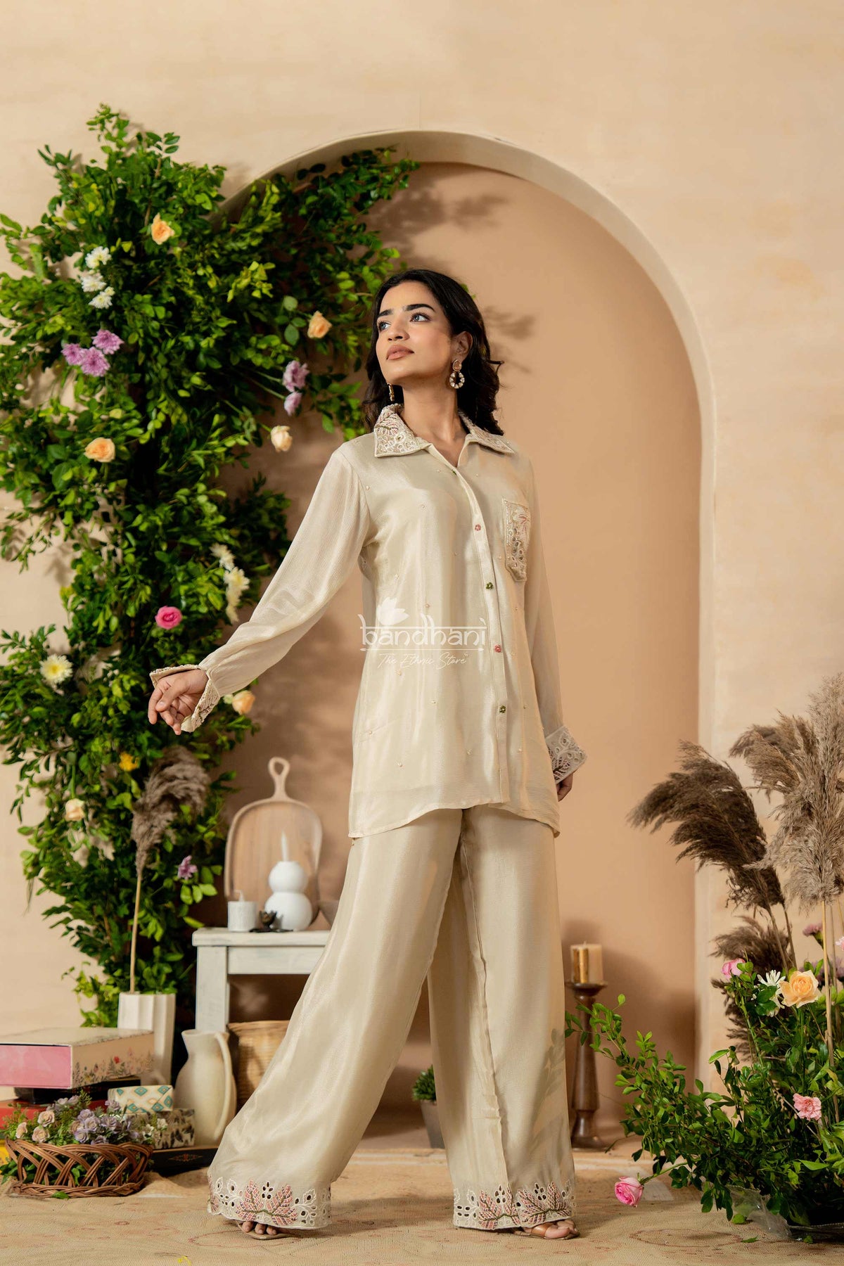 Woman in a beige traditional outfit standing in a decorated indoor setting with plants and flowers.