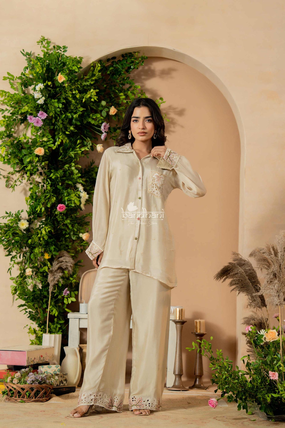 Woman in a beige traditional outfit standing in a decorated indoor setting with flowers and greenery.