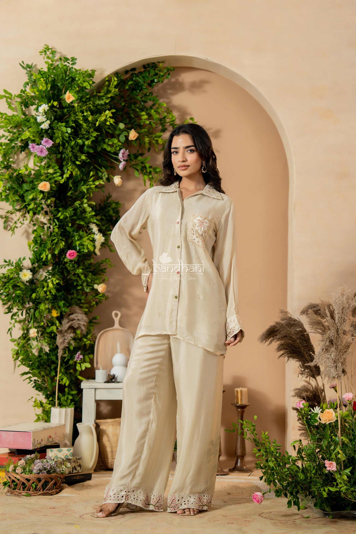 Woman in a beige outfit standing in a decorated room with plants and flowers.