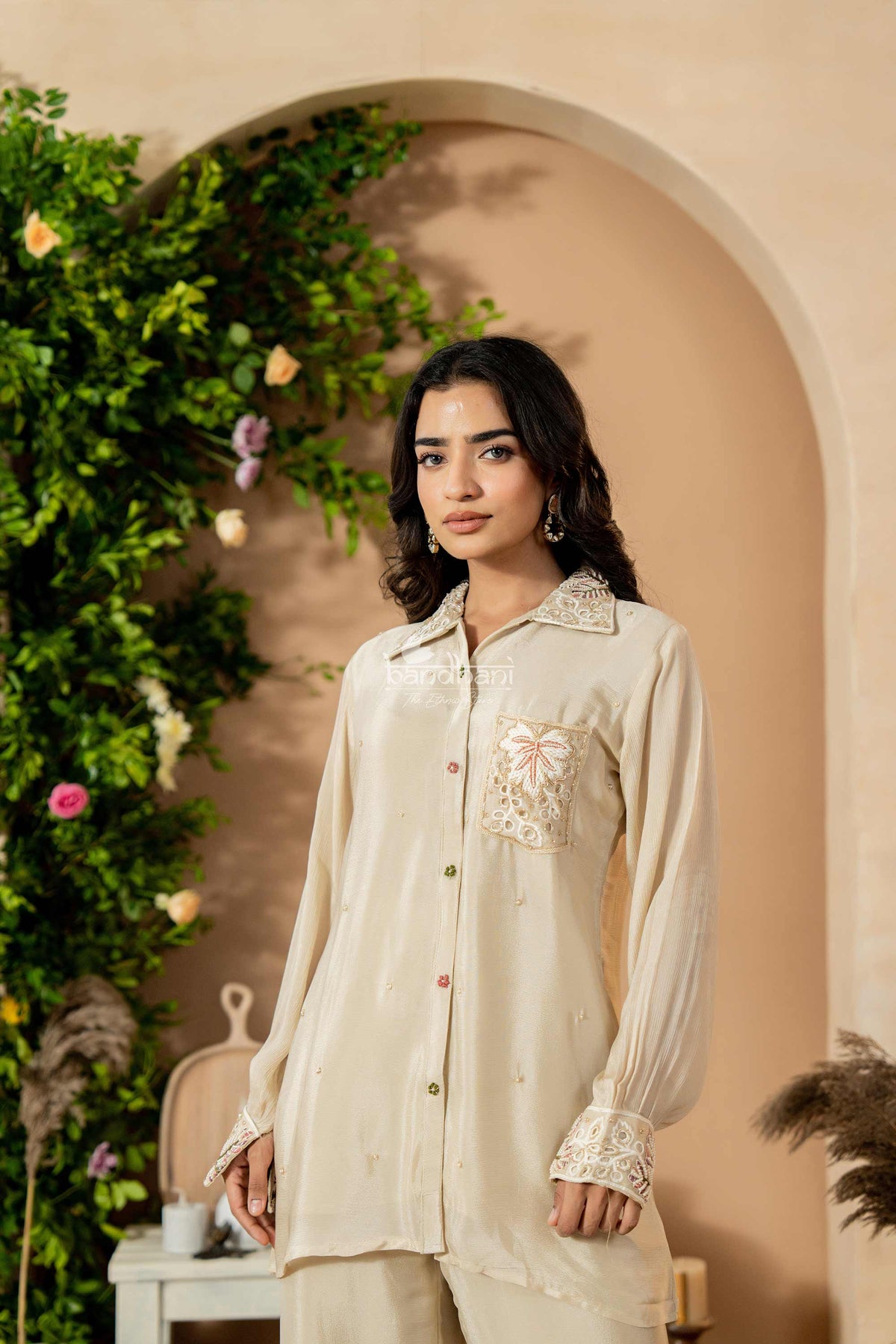 Woman wearing a cream-colored traditional outfit with floral embroidery in a decorative setting.
