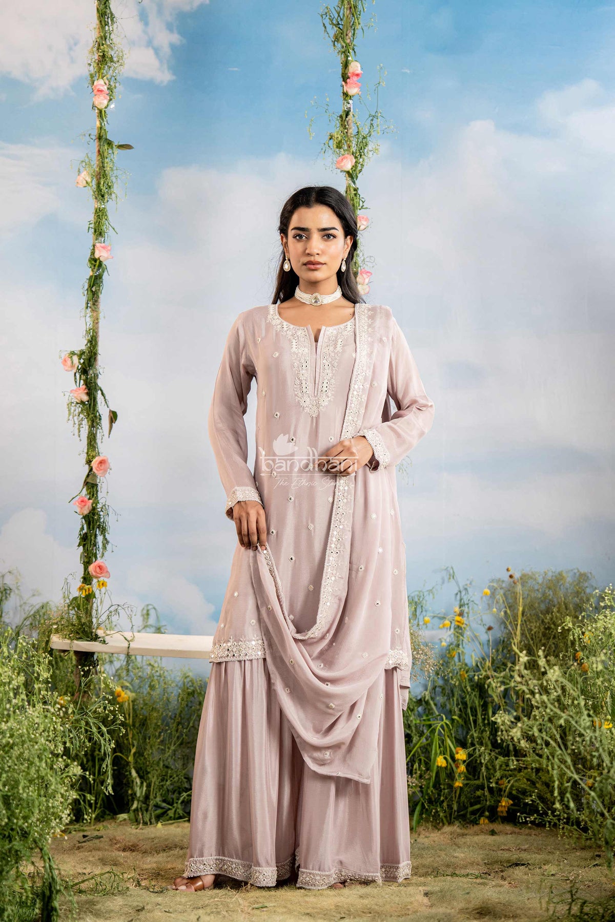 Woman in a light pink traditional outfit standing outdoors with a swing and flowers in the background