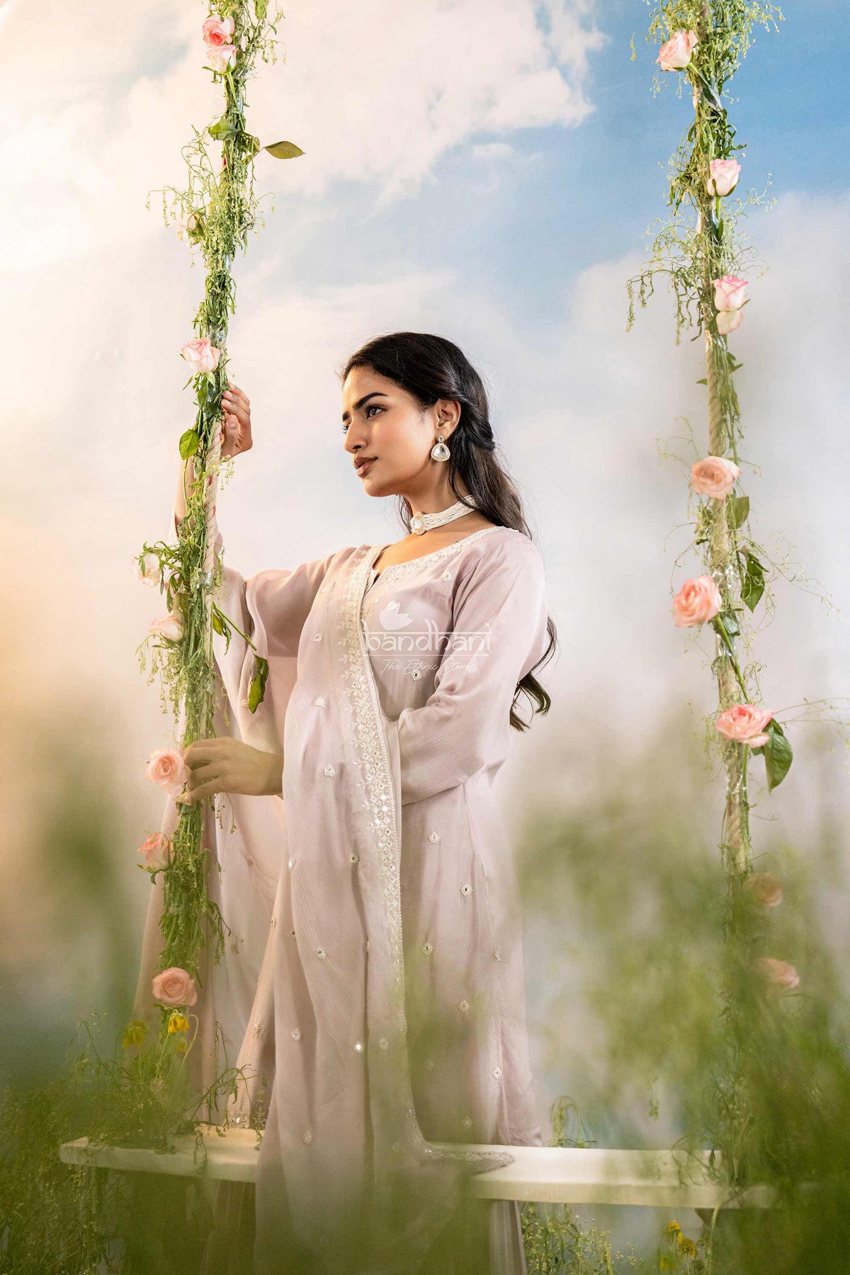 Woman in a light pink traditional outfit sitting on a swing decorated with flowers against a blue sky.
