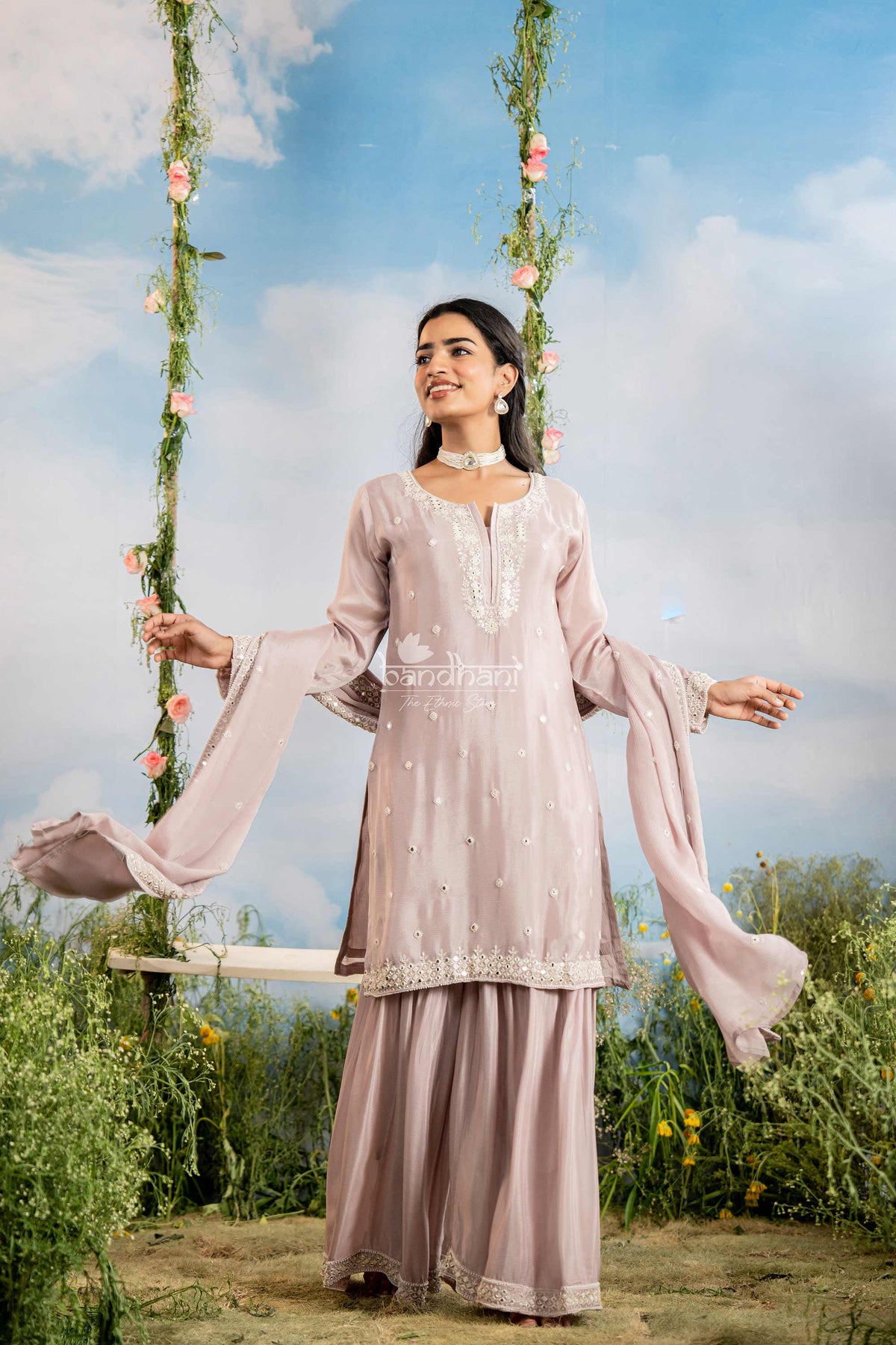 Woman in a light pink traditional outfit sitting on a swing with floral decorations against a blue sky.