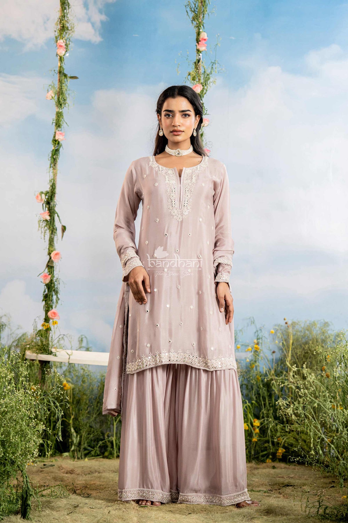 Woman in a light pink traditional outfit standing outdoors with floral decorations and a blue sky.