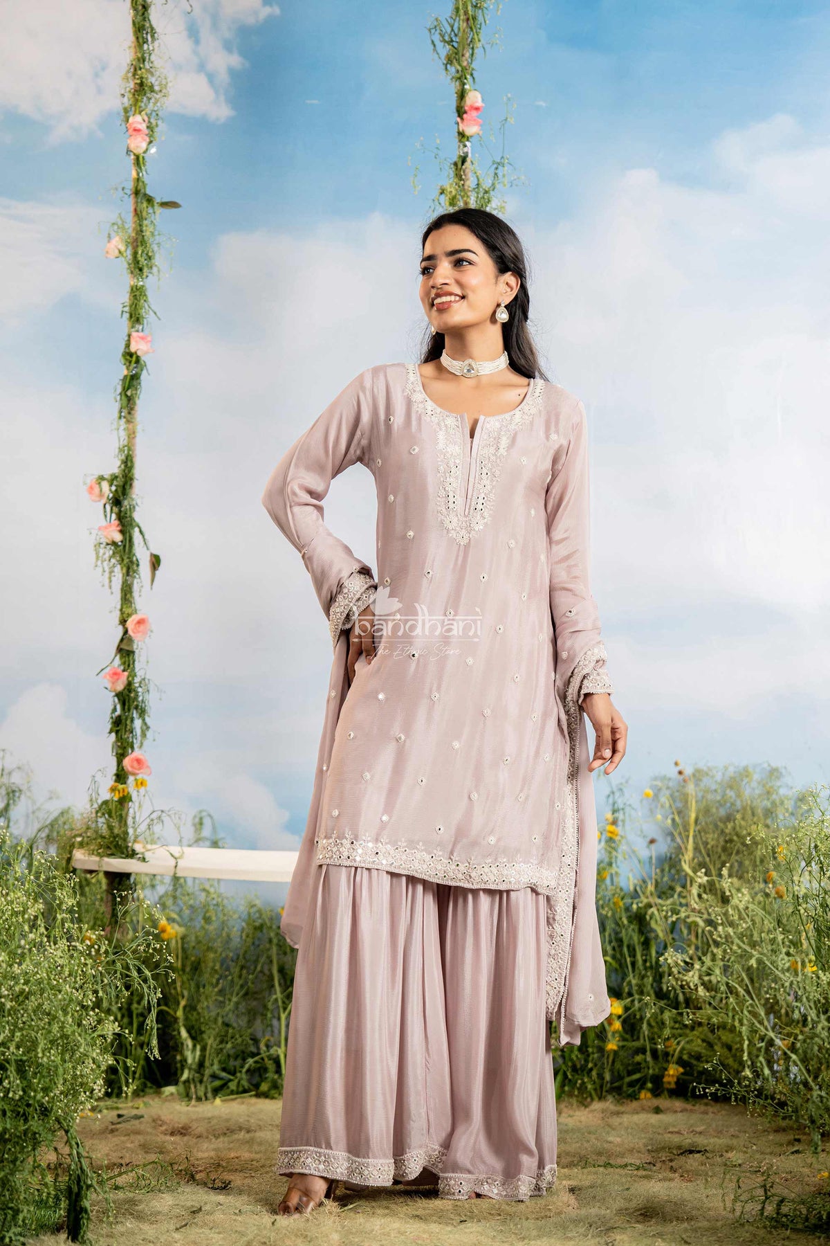 Woman in a light pink traditional outfit standing outdoors with a swing and floral decorations in the background.