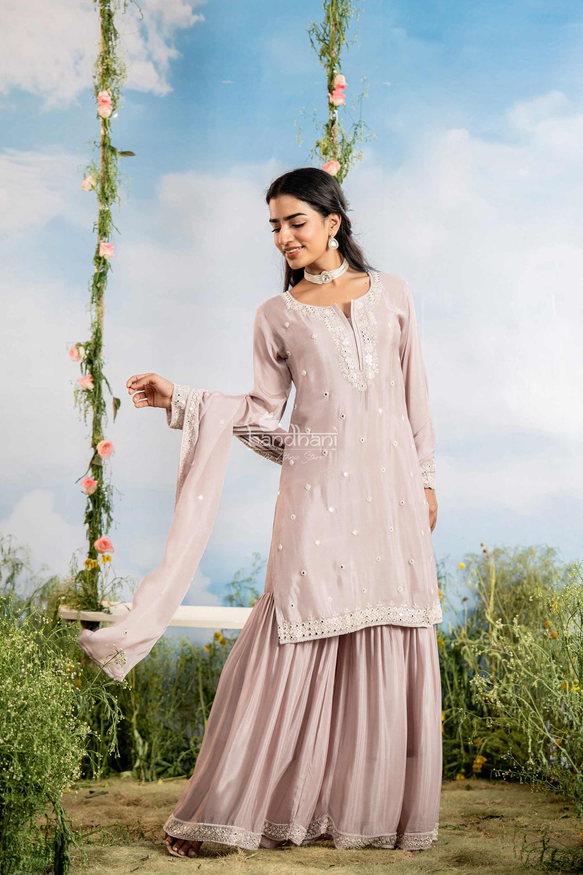 Woman in a light pink traditional outfit sitting on a swing with floral decorations against a blue sky.