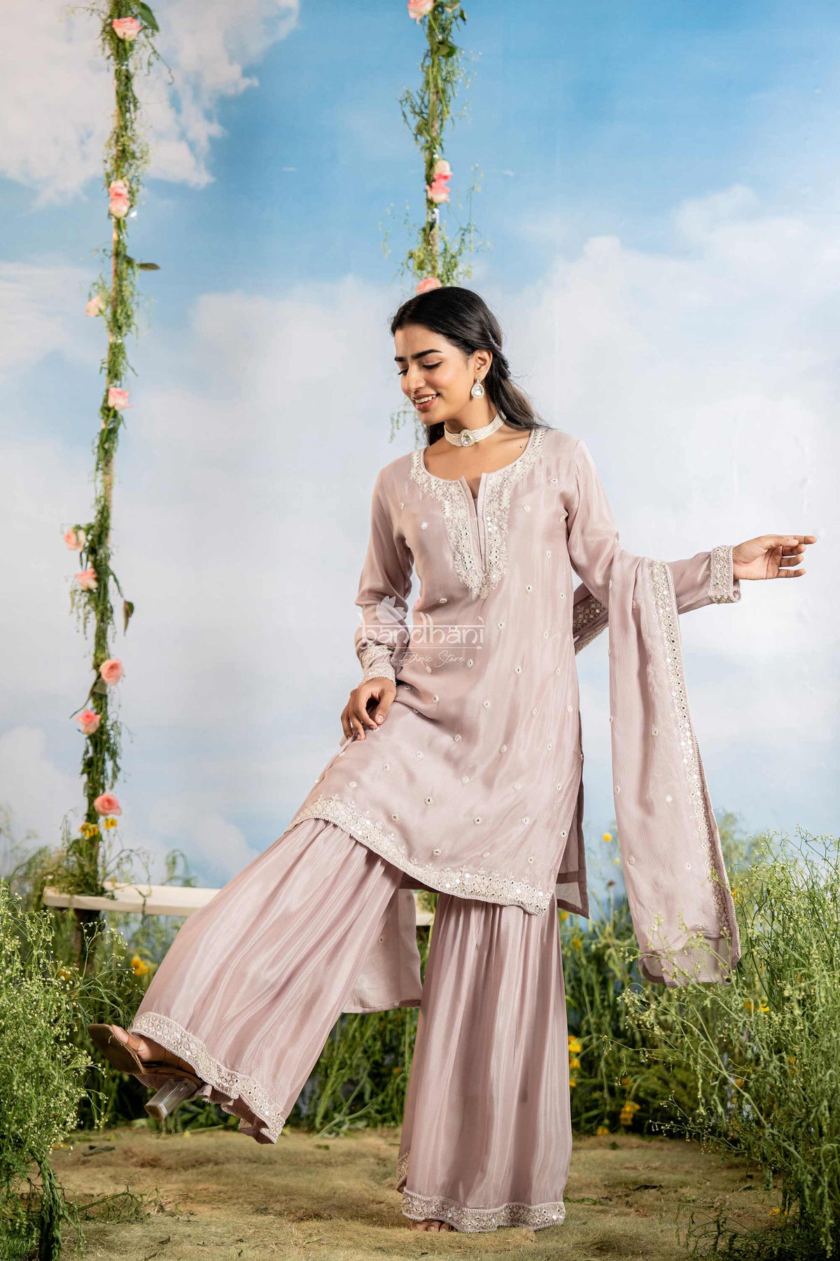 Woman in a light pink traditional outfit standing outdoors with a floral swing and blue sky in the background