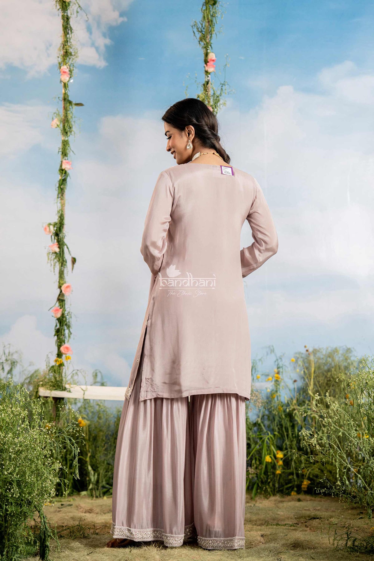 Woman in a light pink traditional outfit standing outdoors with greenery and a blue sky.