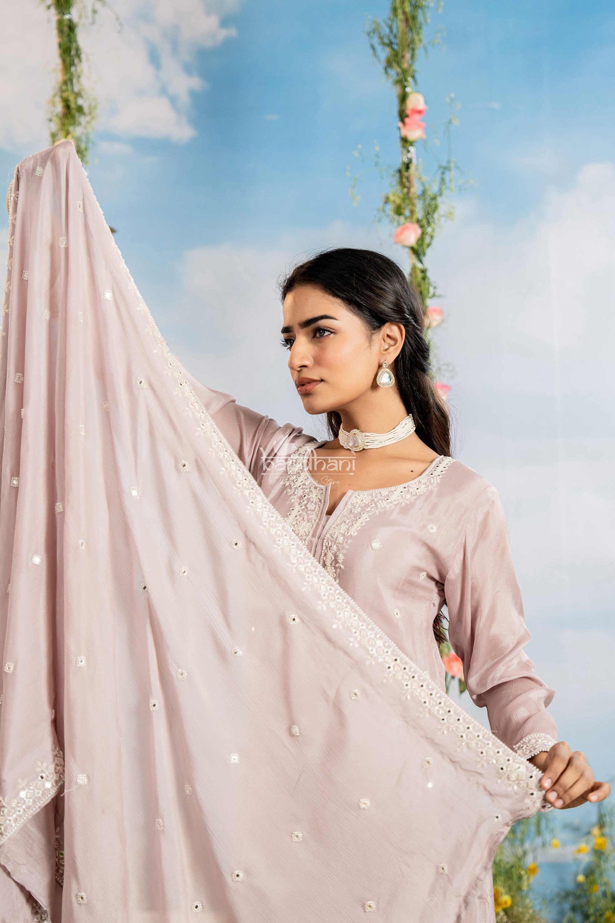 Woman holding a light pink dupatta with floral decorations against a blue sky.