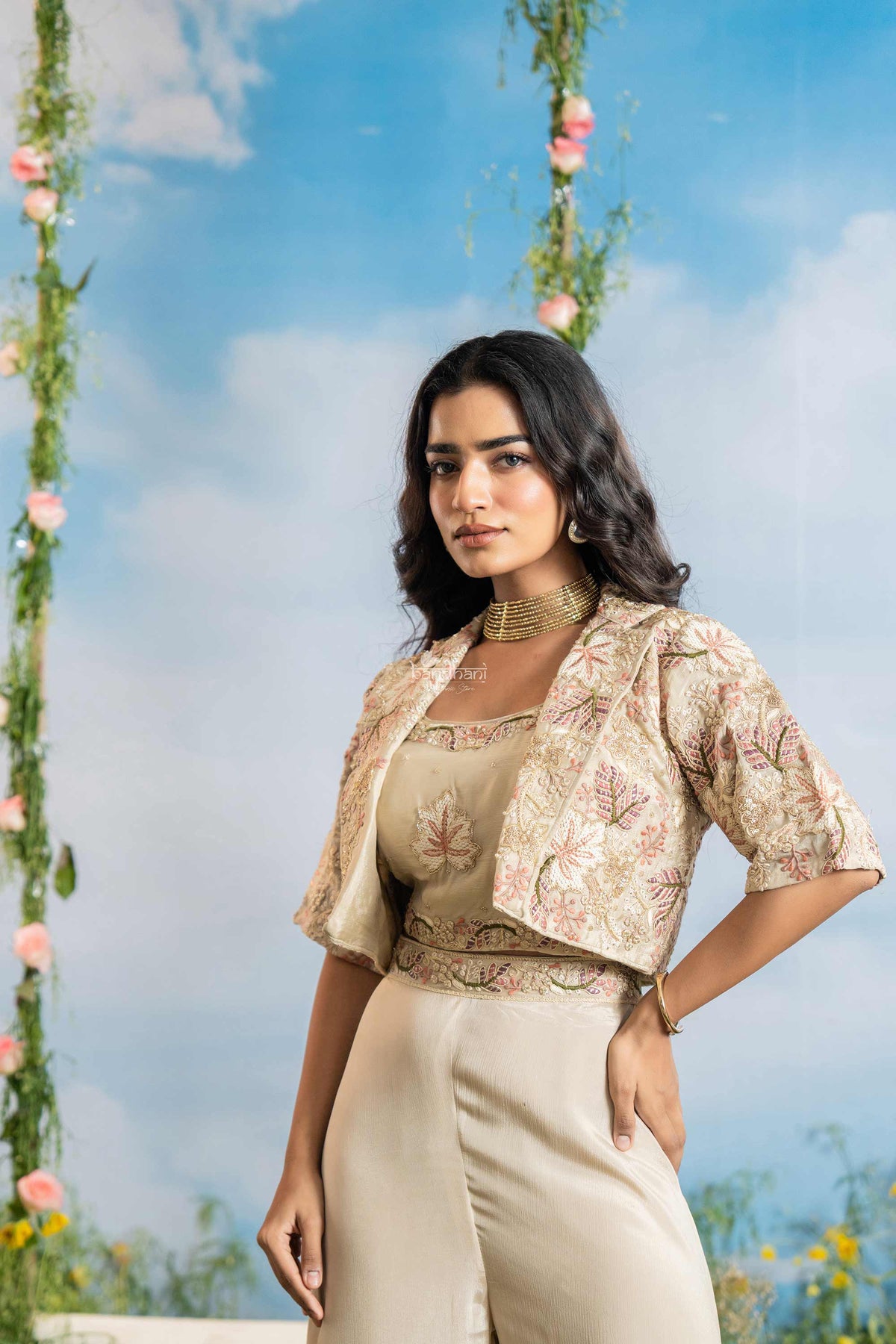 Woman in a patterned top and beige pants standing against a blue sky with floral decorations.