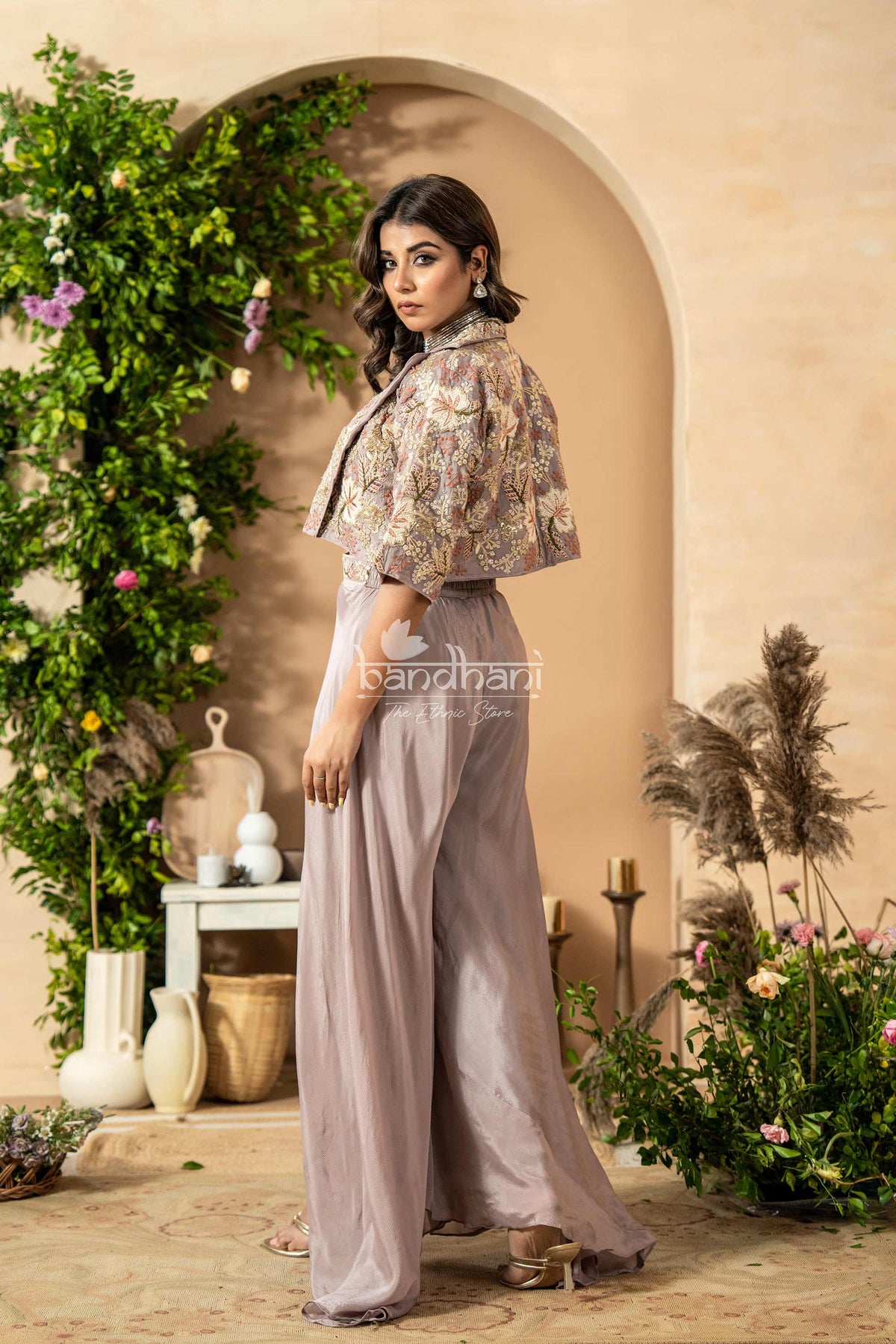 Woman in a floral top and light gray pants standing in a decorated outdoor setting with plants and archway.