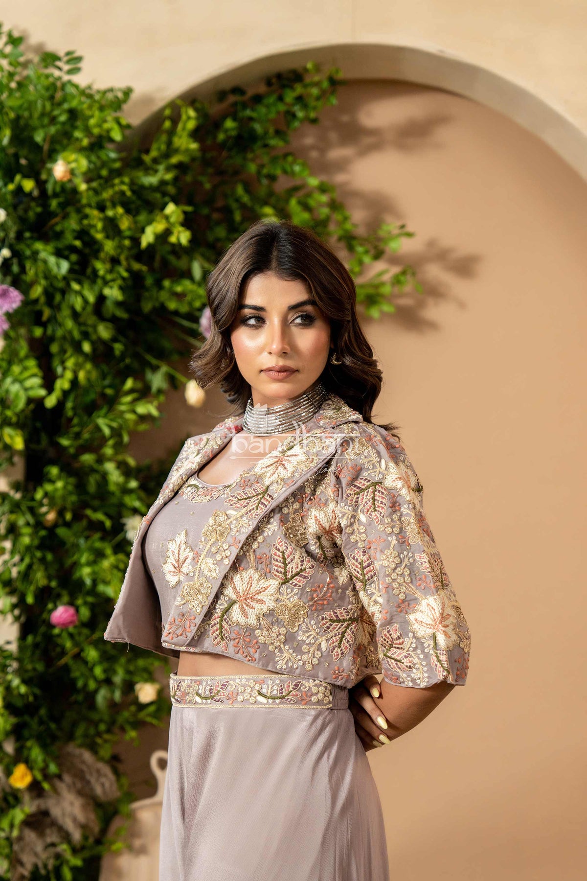 Woman wearing a floral embroidered jacket and gray dress against a beige wall with greenery.