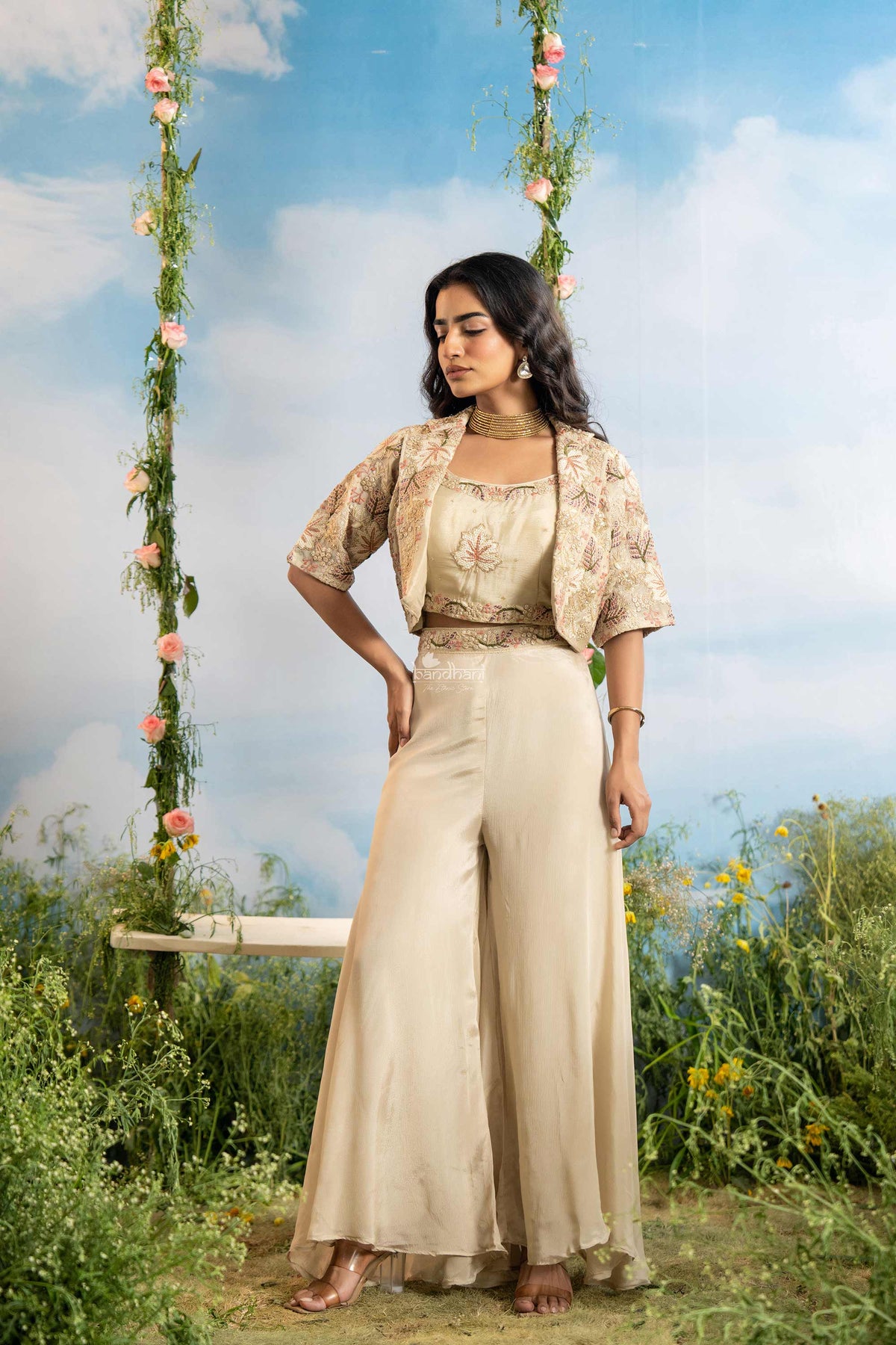Woman in a beige outfit standing next to a floral swing with a blue sky and greenery in the background