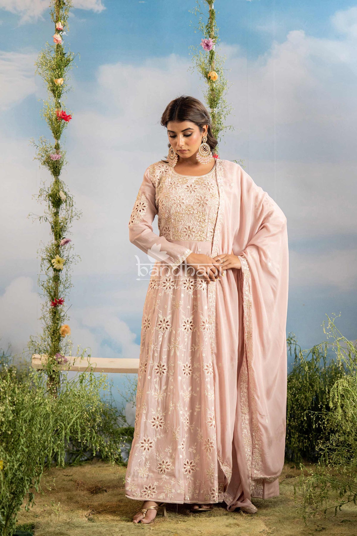 Woman in a light pink traditional outfit standing outdoors with greenery and a blue sky in the background.
