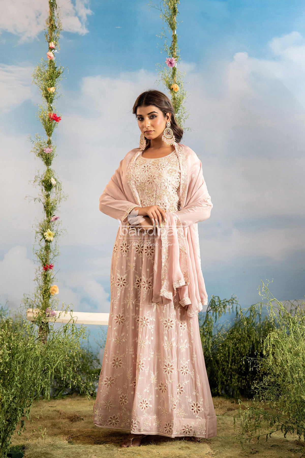Woman in a light pink traditional outfit sitting on a swing with floral decorations against a blue sky.