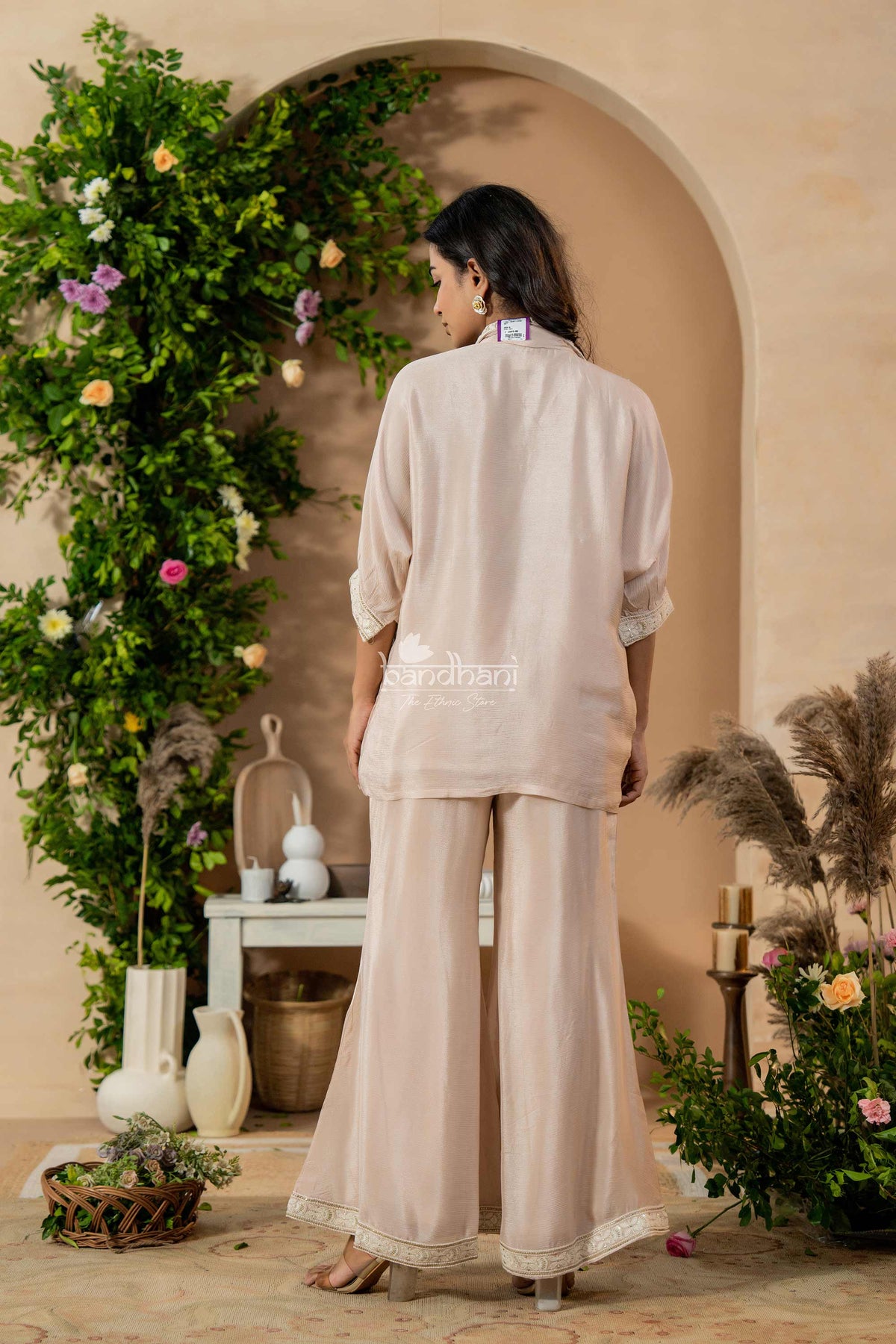 Woman in a beige outfit standing in a decorated room with flowers and plants.