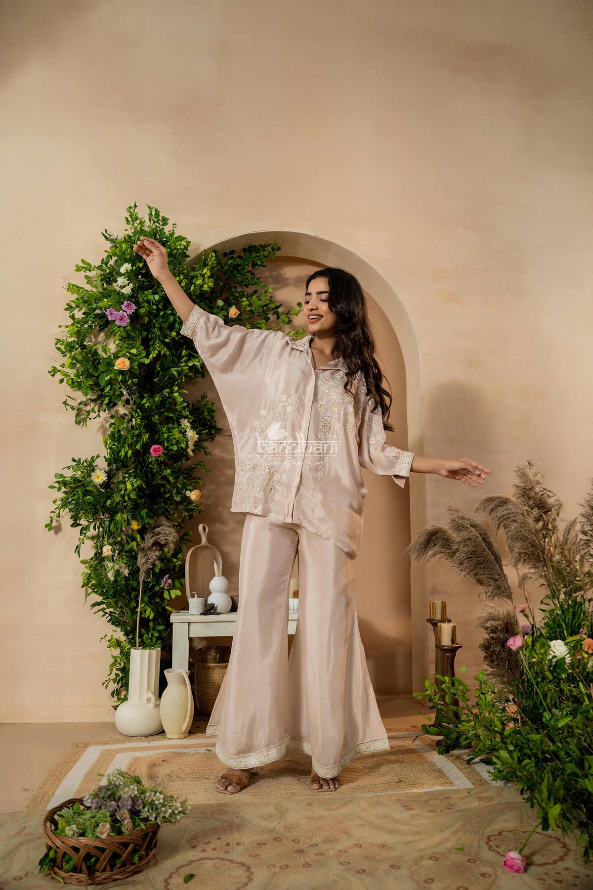 Woman in a beige outfit standing in a decorated room with plants and a table.