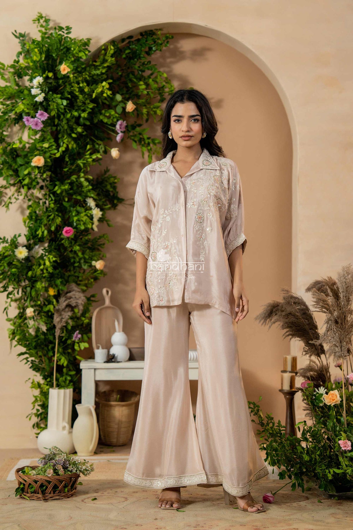 Woman in a beige outfit standing in a decorated room with plants and flowers.