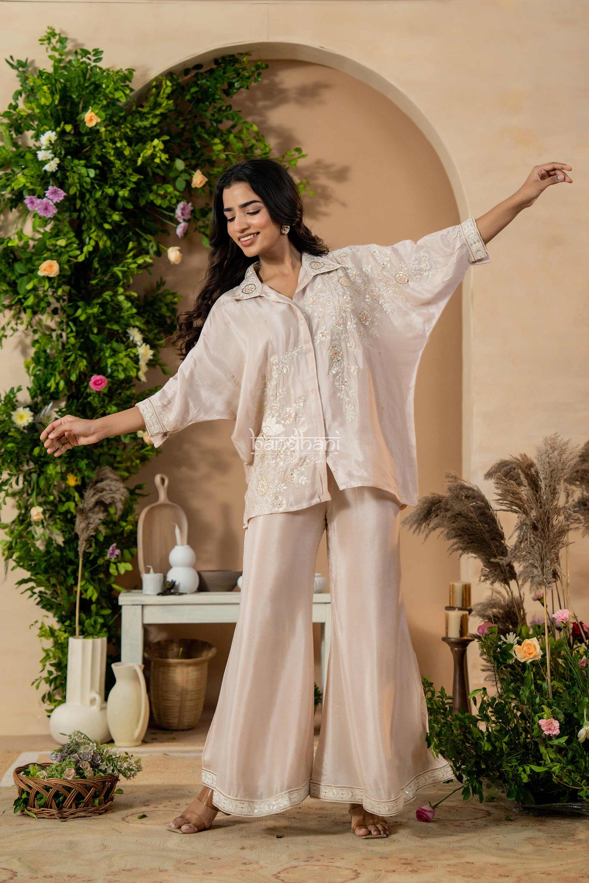 Woman in a beige outfit standing in a decorated room with plants and a vase.