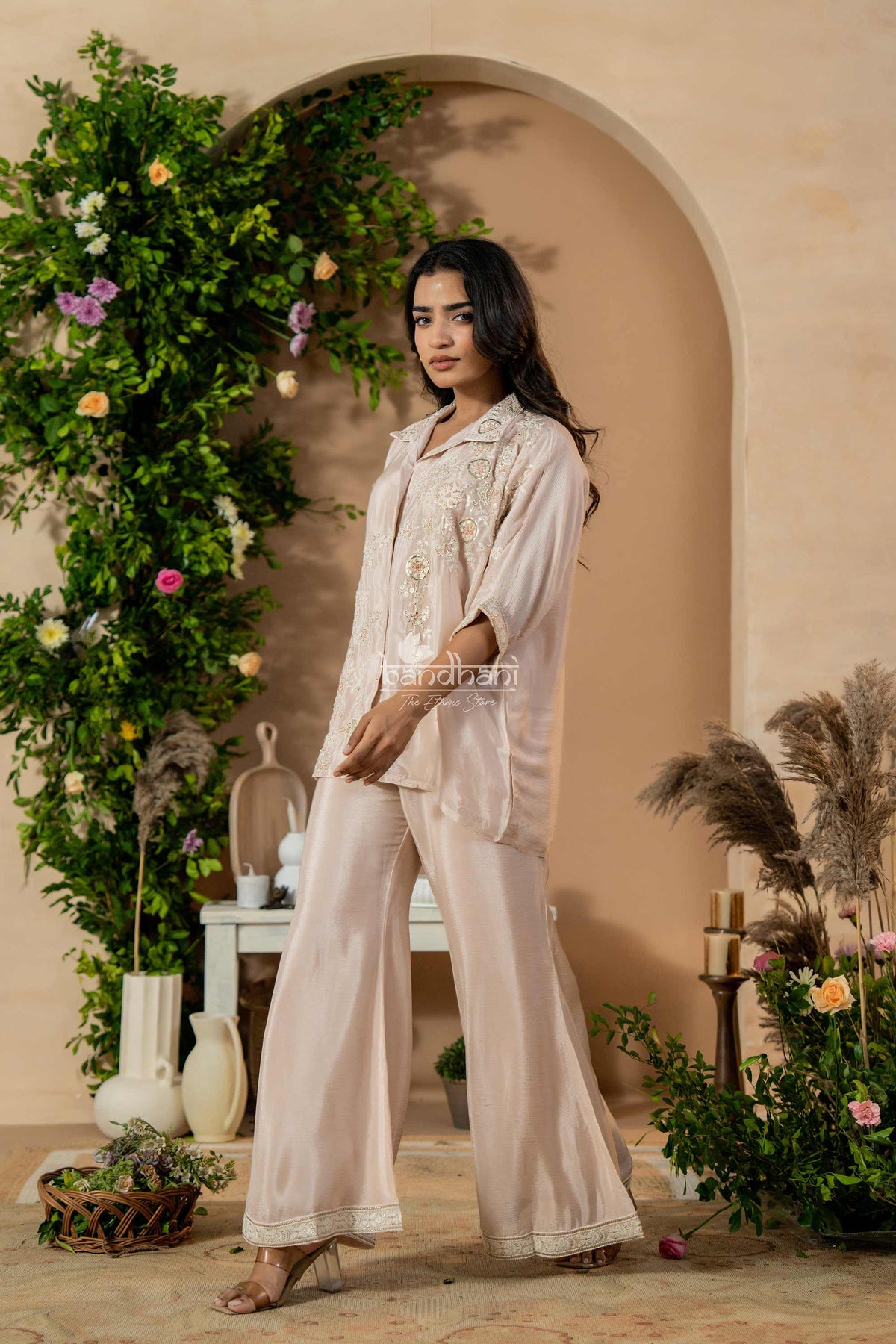Woman in a beige outfit standing in a decorated indoor setting with plants and flowers.