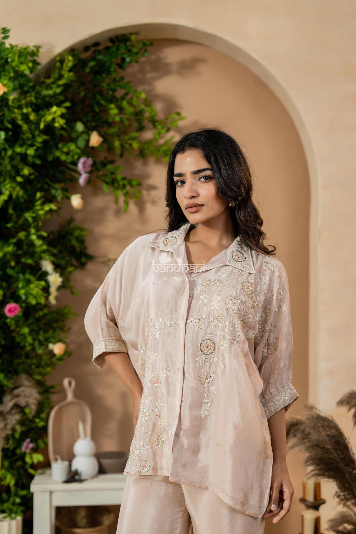 Woman wearing a beige embroidered outfit standing in a decorative indoor setting with plants and a table.