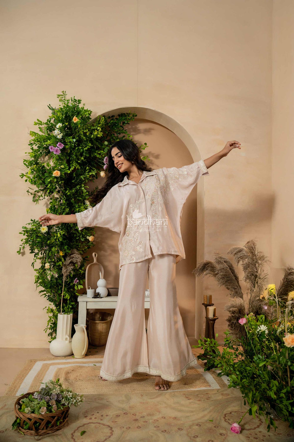 Woman in a beige outfit standing in a decorated room with plants and flowers.