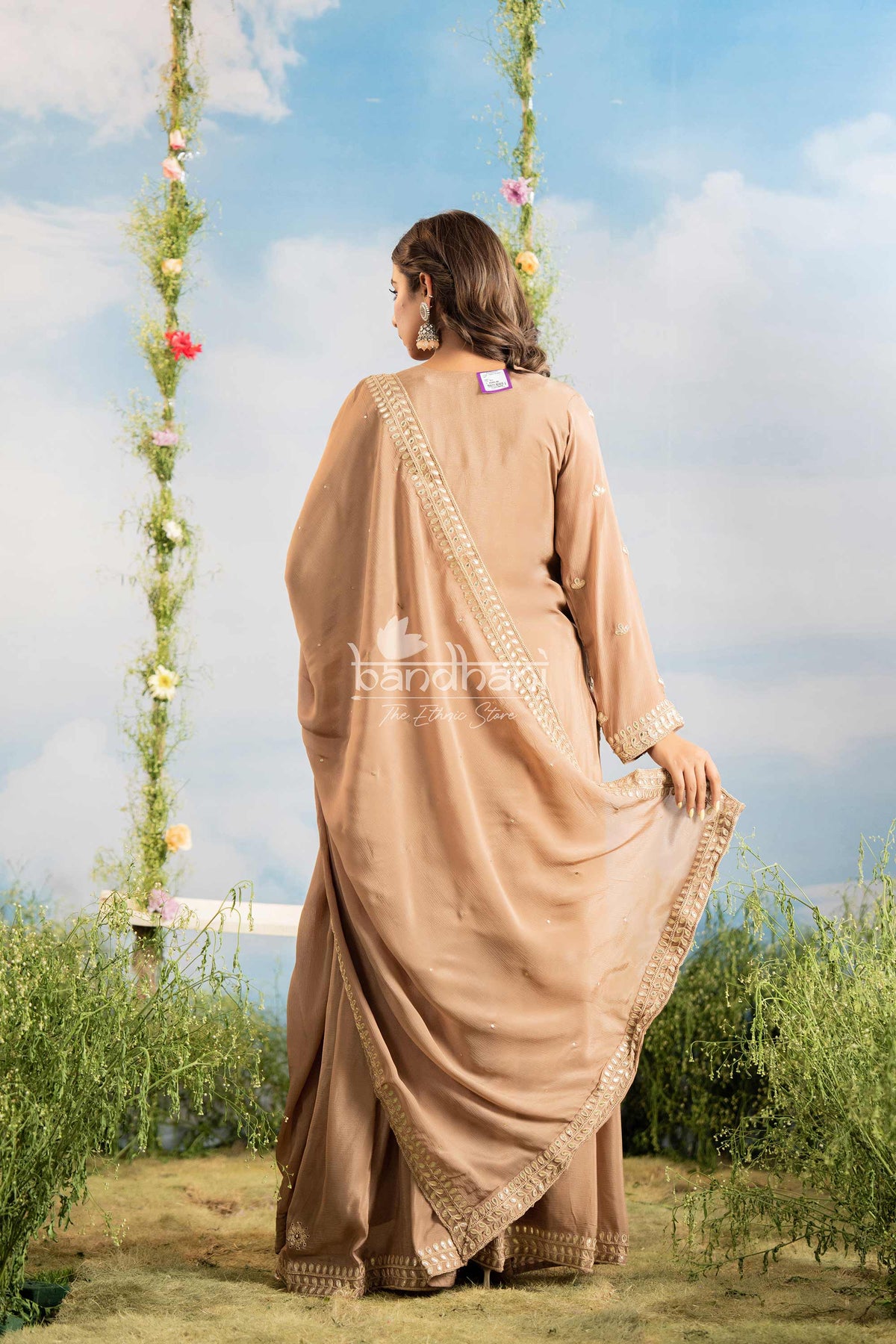 Woman in a beige saree standing outdoors with greenery and a blue sky in the background