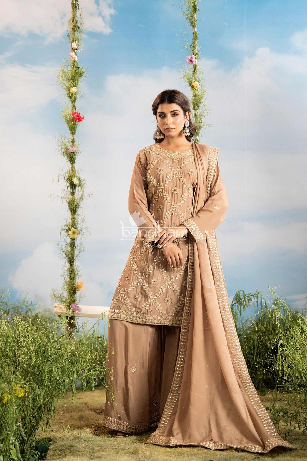 Woman in a beige traditional outfit standing outdoors with greenery and flowers in the background
