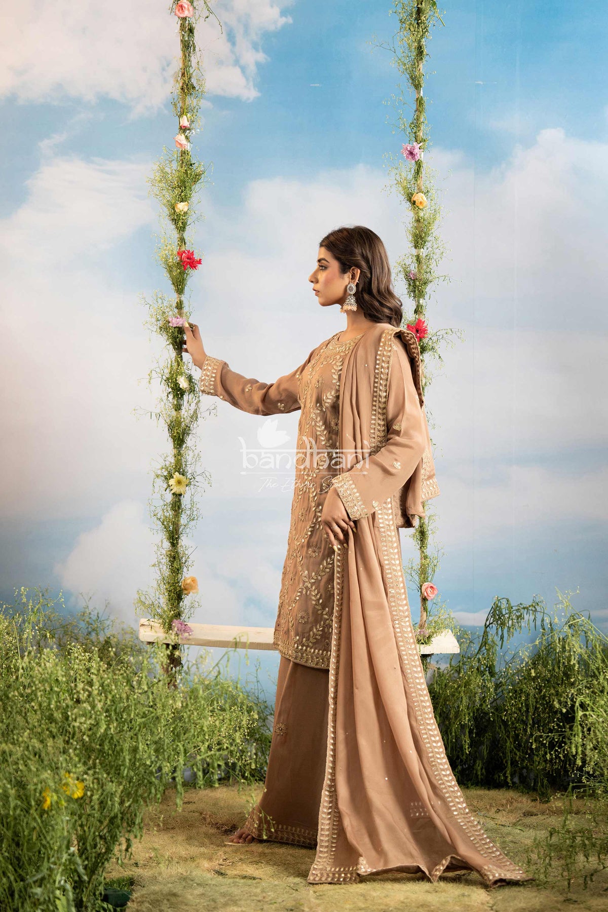 Woman in a beige traditional outfit sitting on a swing with floral decorations against a blue sky.