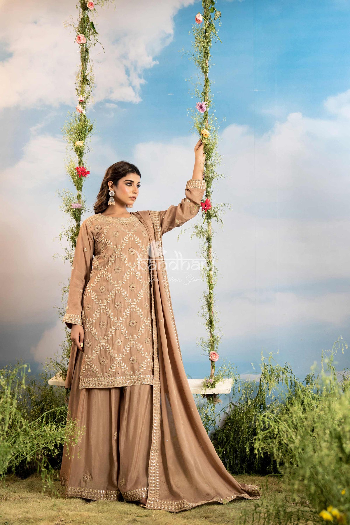 Woman in a beige traditional outfit sitting on a swing with floral decorations against a blue sky.