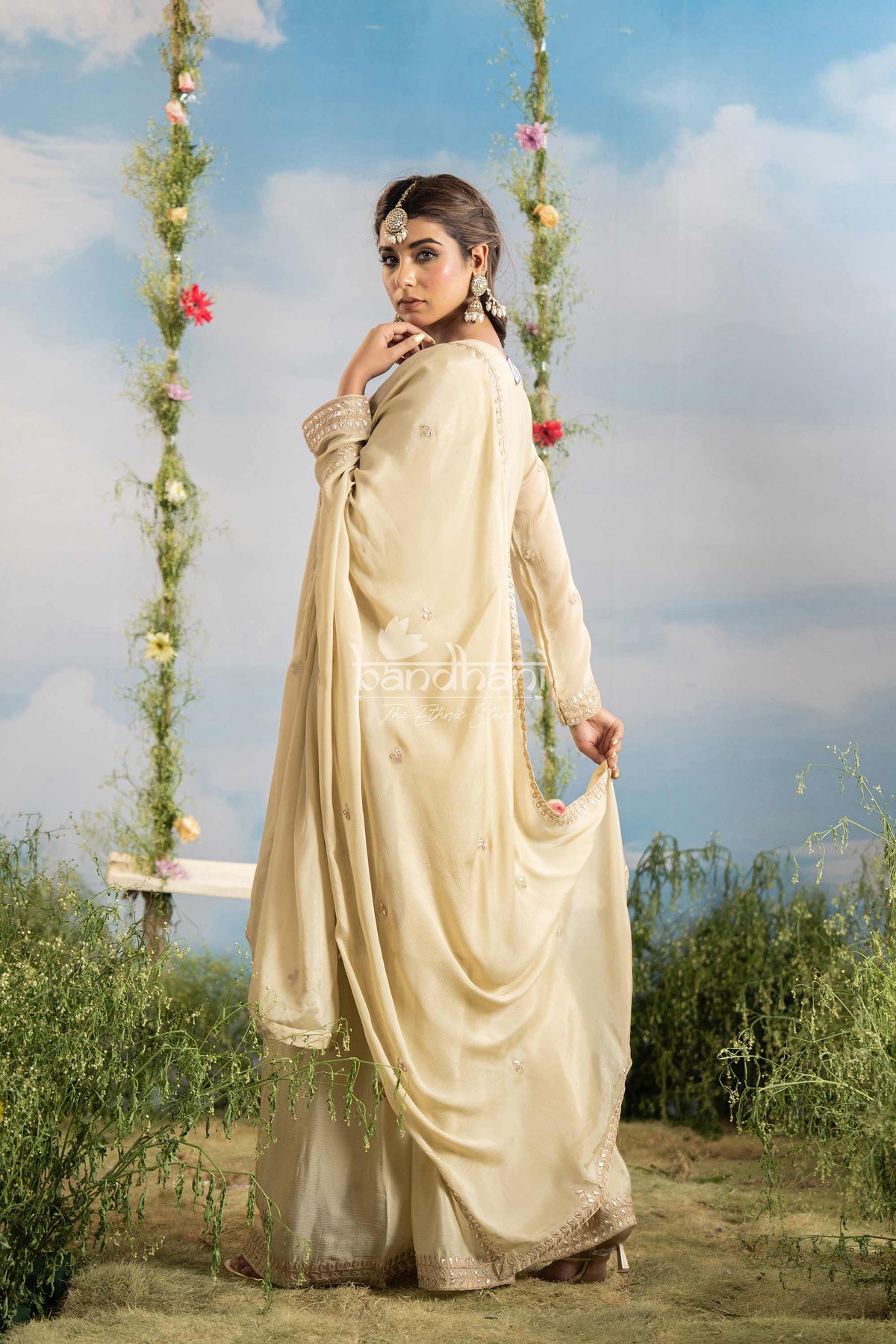 Woman in a beige saree standing outdoors with greenery and a blue sky in the background