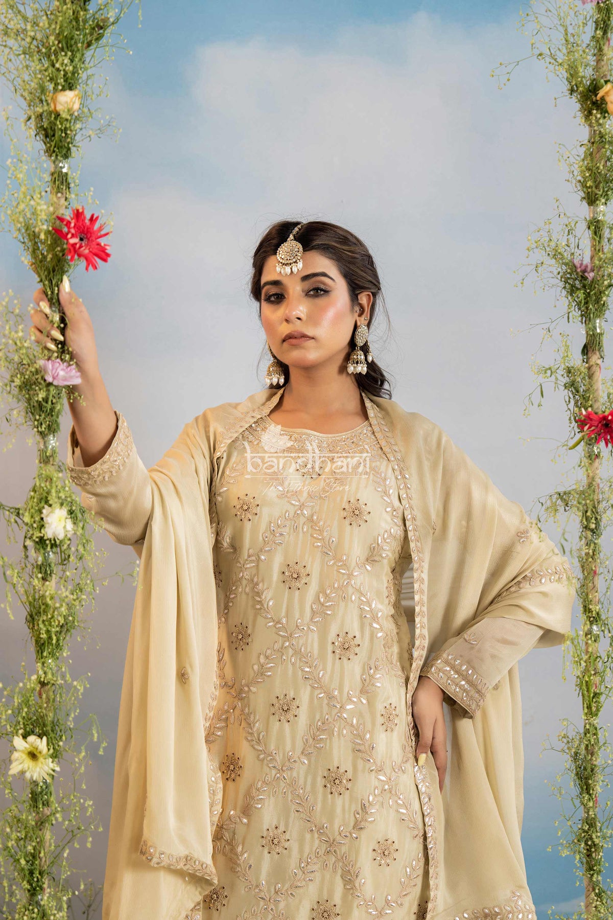 Woman in a beige embroidered outfit holding flowers against a blue sky background