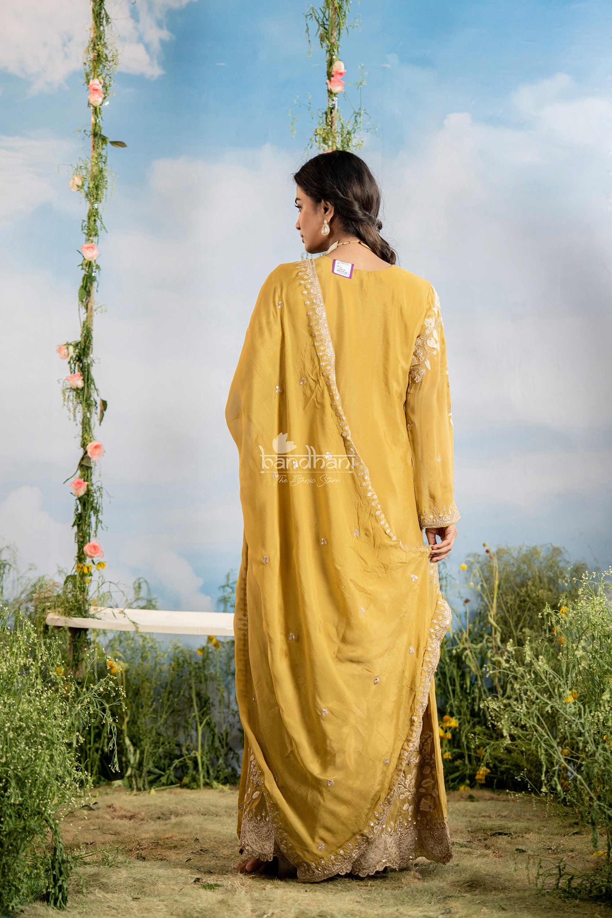 Woman in a yellow traditional outfit standing outdoors with greenery and a blue sky in the background.