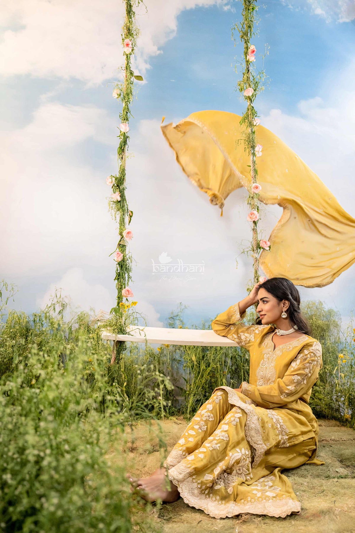 Woman in a yellow traditional outfit sitting on a swing with floral decorations against a blue sky.