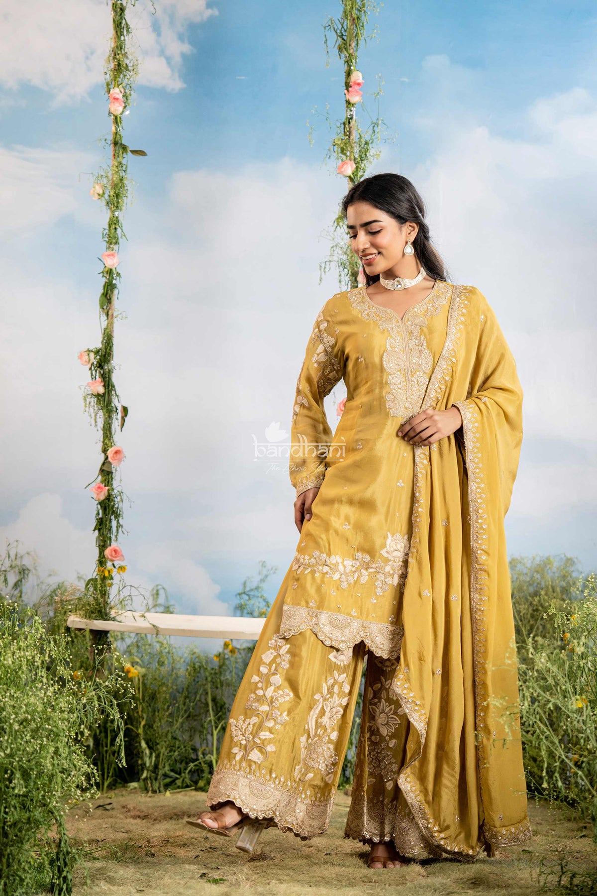 Woman in a yellow traditional outfit standing outdoors with greenery and a blue sky.