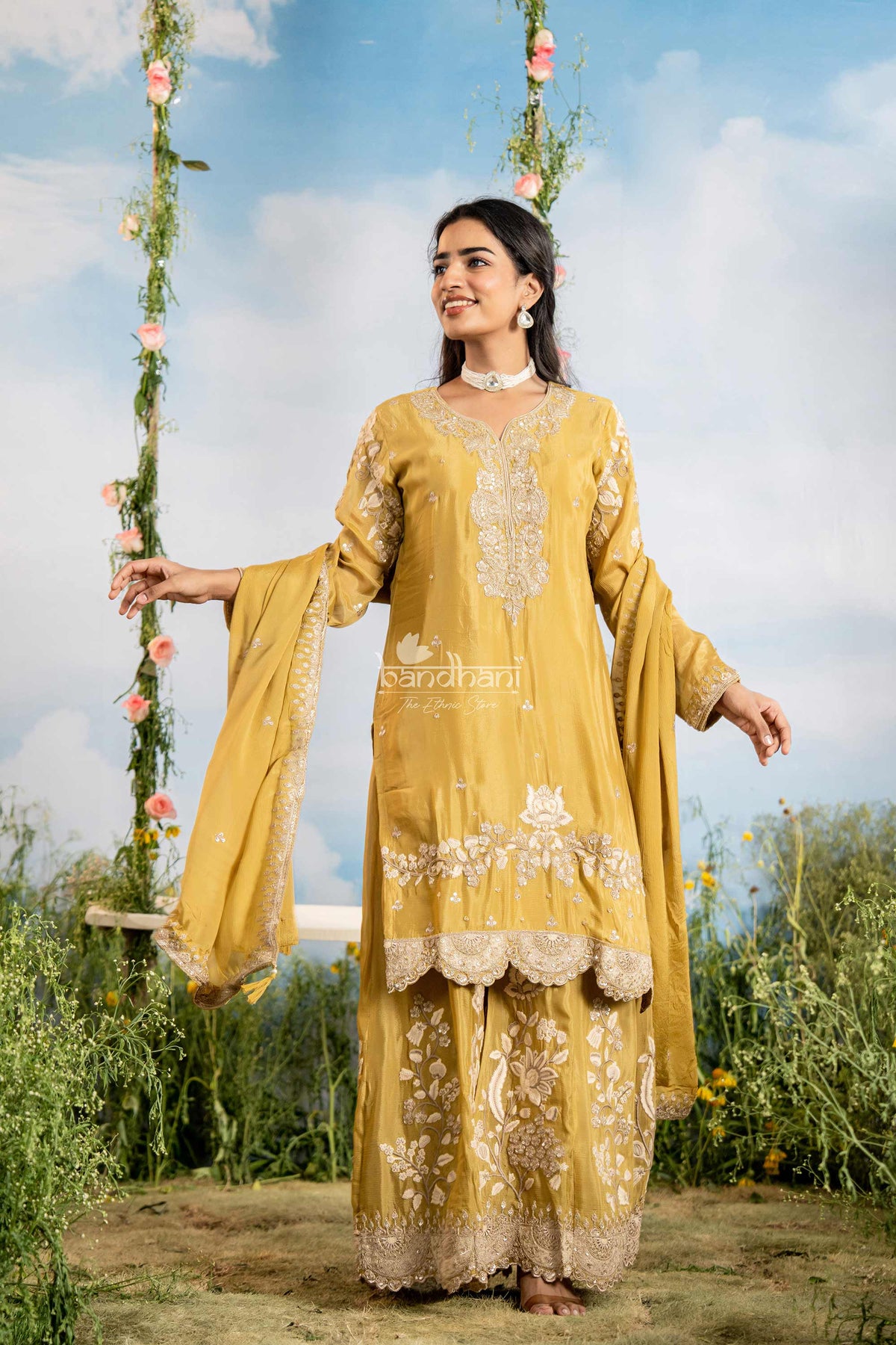 Woman in a yellow embroidered outfit sitting on a swing with floral decorations against a blue sky.