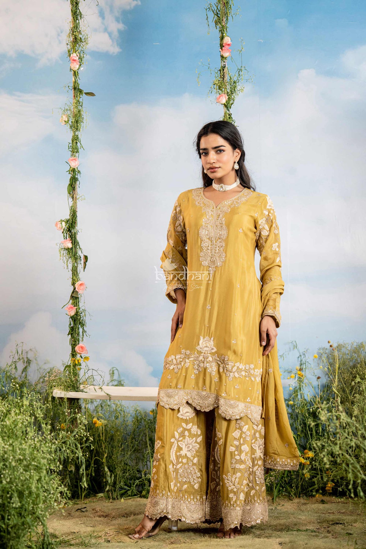 Woman in a yellow traditional outfit standing on a swing with floral decorations against a blue sky.
