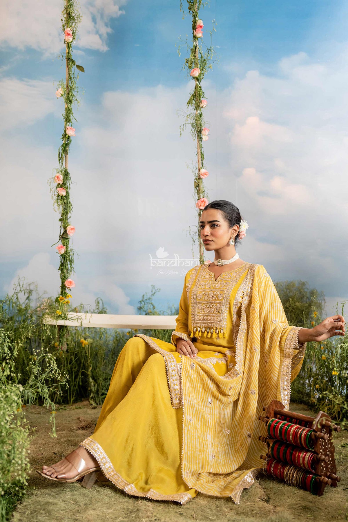 Woman in a yellow traditional outfit sitting on a swing with floral decorations against a blue sky.