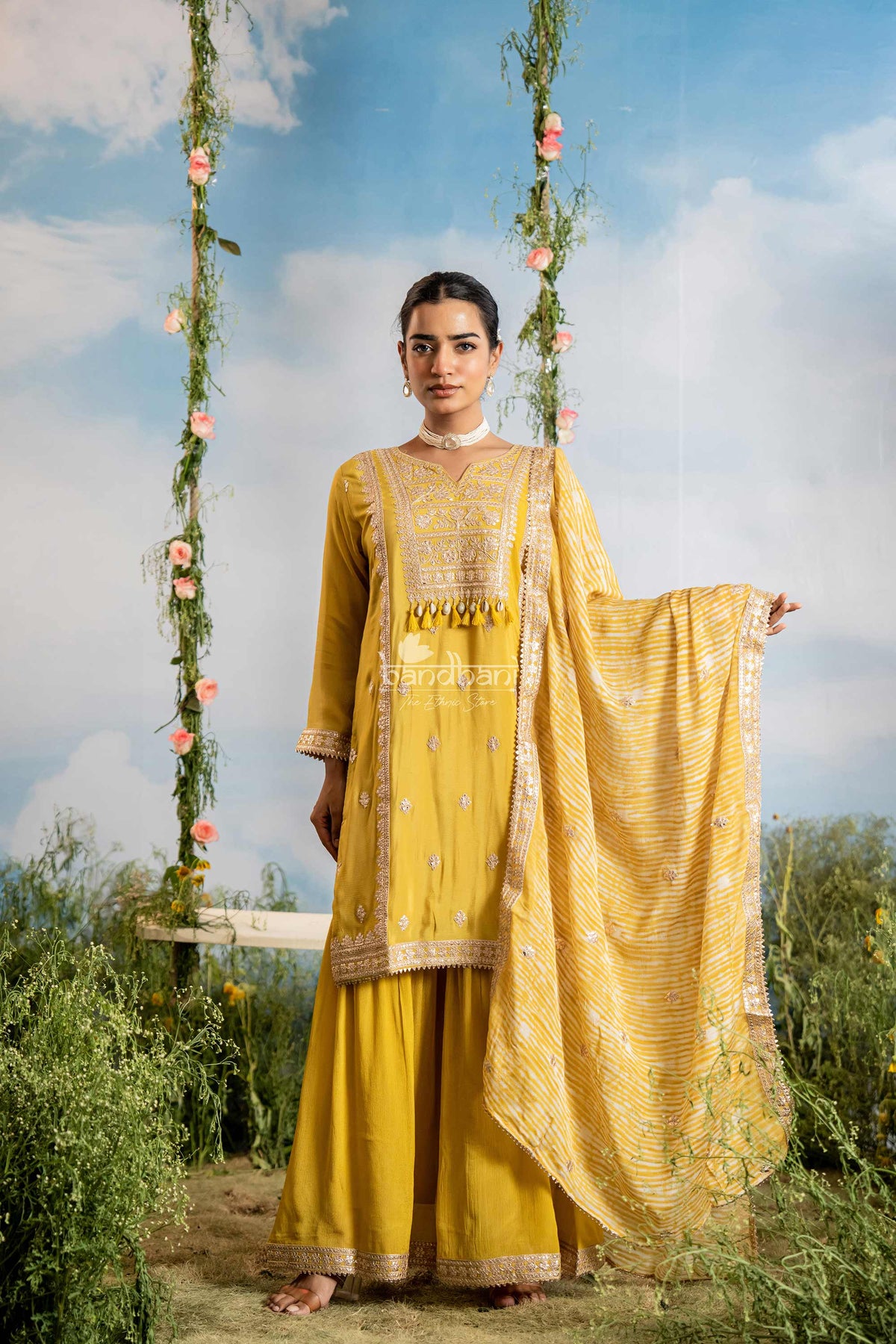 Woman in a yellow traditional outfit standing outdoors with a swing and floral decorations in the background.