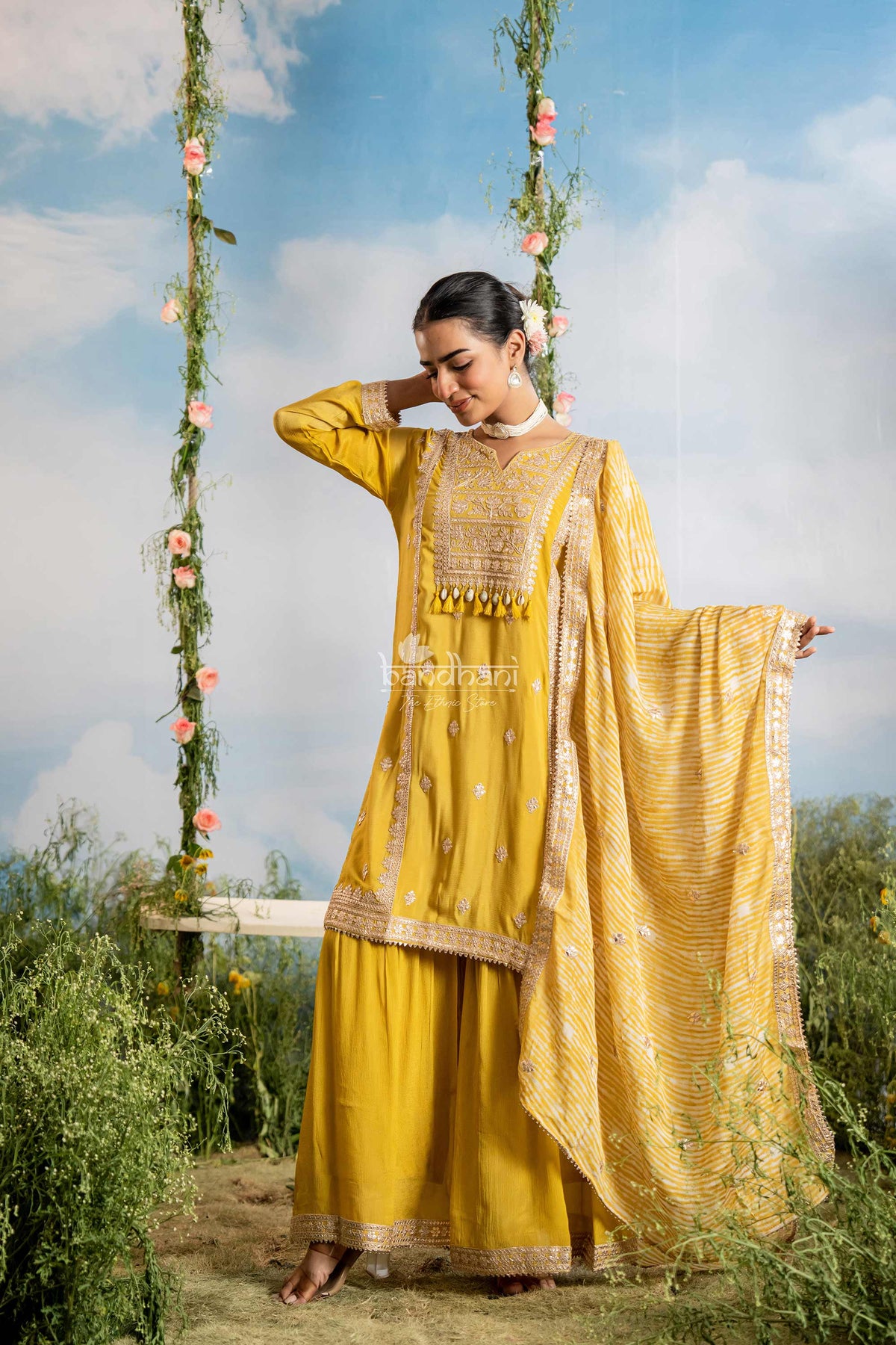 Woman in a yellow traditional outfit sitting on a swing with floral decorations against a blue sky.