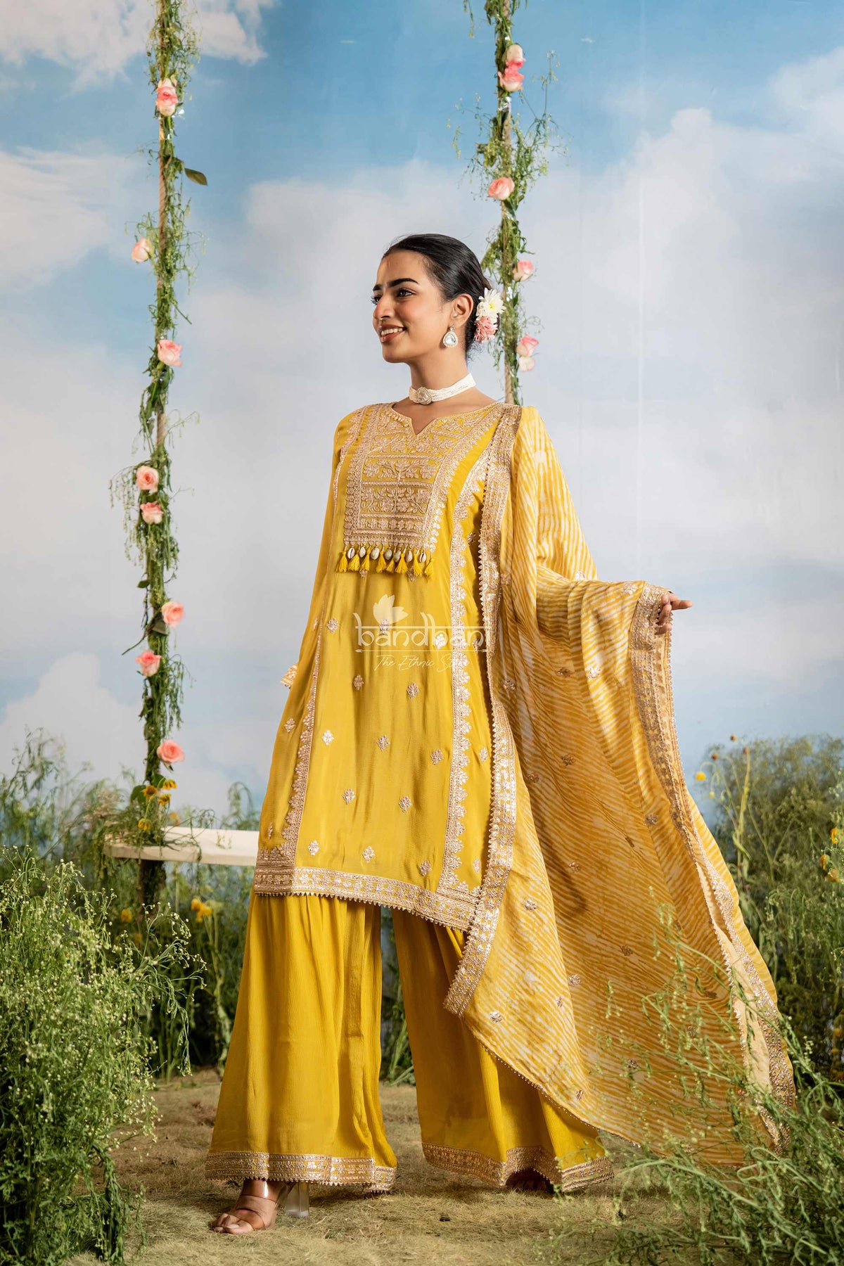 Woman in a yellow traditional outfit sitting on a swing with floral decorations against a blue sky.